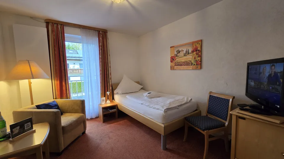 Einzelzimmer mit Einzelbett, Sessel, Fernseher und Balkontür
