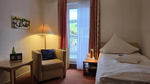 Einzelzimmer mit Sessel und Stehlampe am Fenster, daneben das Einzelbett